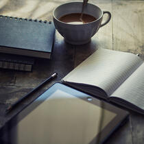 Photo of coffee cup, notebooks, electronic tablet, and pencil on a table